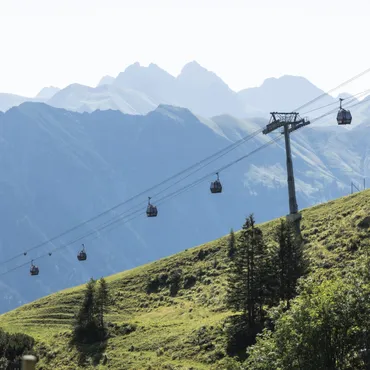 Herbst-Online-Buchungs-Special Seilbahn fährt einen grünen Almenhang hinauf mit Berggipfeln im Hintergrund